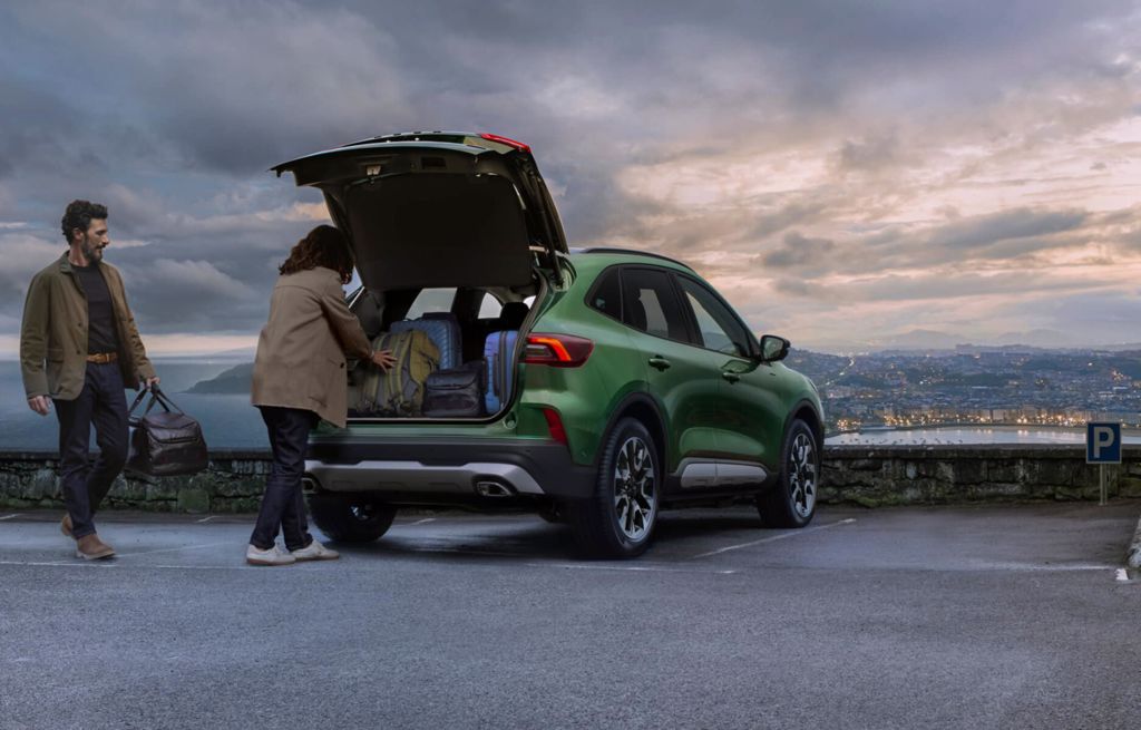 Kobieta pakująca bagaż do zielonego Forda Kuga 2.5 FHEV ST-Line X na tle widoku miasta.